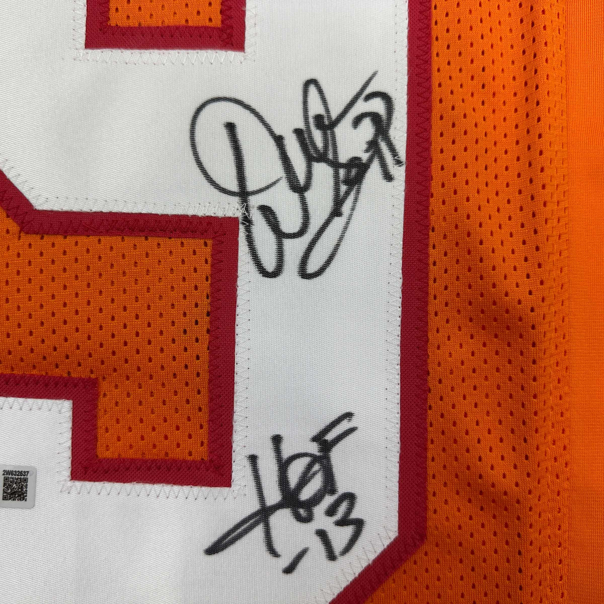 Close-up of orange mesh jersey fabric with a red-edged white number panel, black marker signatures, and a small authentication tag at lower left.