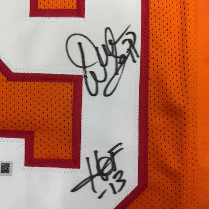Close-up of orange mesh jersey fabric with a red-edged white number panel, black marker signatures, and a small authentication tag at lower left.