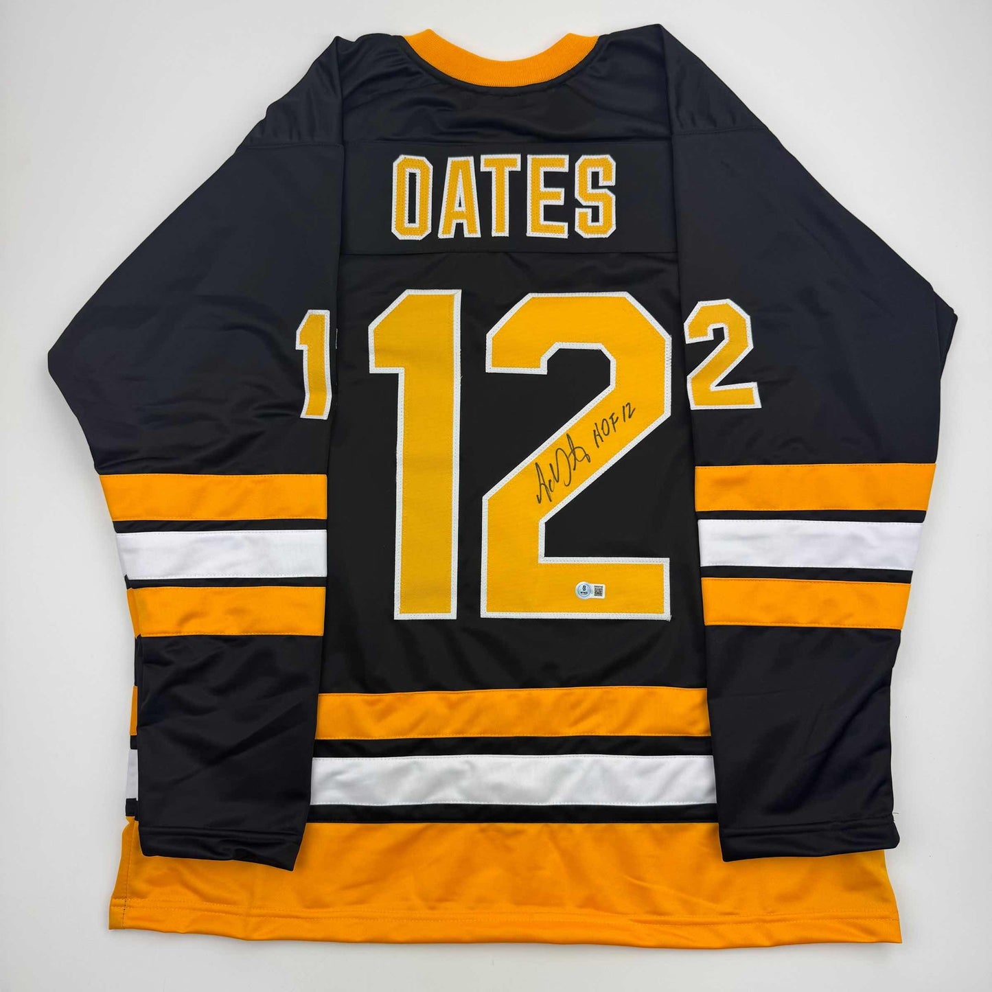 Back view of black and orange striped Boston hockey jersey numbered 12 with "OATES" and handwritten "HOF 12" inscription.