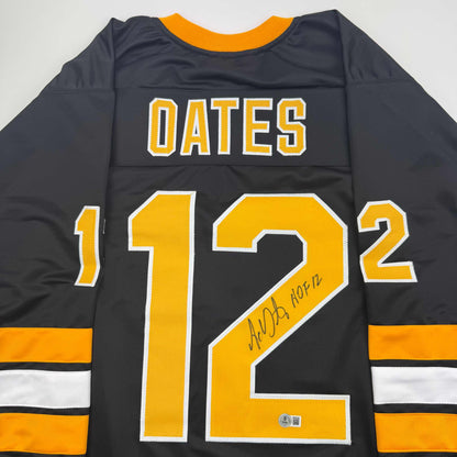 Back view of black Boston Bruins hockey jersey displaying "OATES" and number "12" in gold with white trim, signed "HOF 12" on the number.
