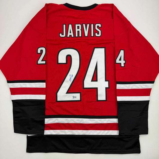Back view of red hockey jersey with "JARVIS" and number "24" in white and black lettering, signed in blue ink, displayed flat against white background.