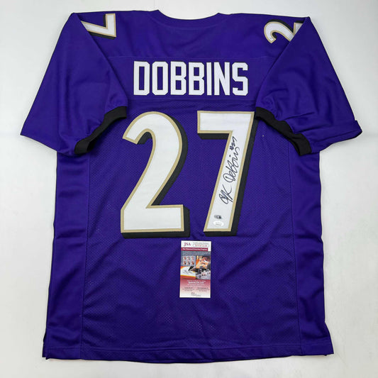 Purple Baltimore Ravens #27 Dobbins football jersey with white and gold numbering displayed flat with JSA authentication certificate visible.