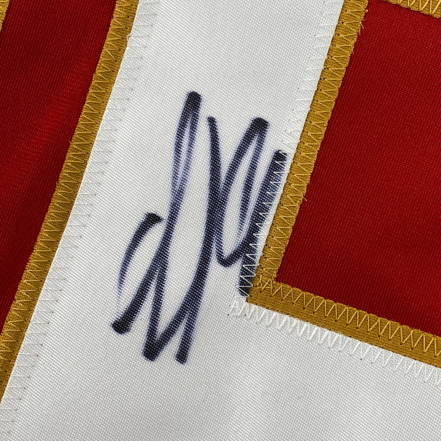 Close-up of handwritten signature in dark ink on white jersey fabric with red and gold trim visible at edges.