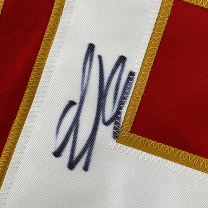 Close-up of handwritten signature in dark ink on white jersey fabric with red and gold trim visible at edges.