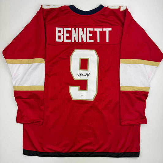 Red hockey jersey with white and gold horizontal stripes, "BENNETT" nameplate, number 9 with signature, displayed flat on white background.