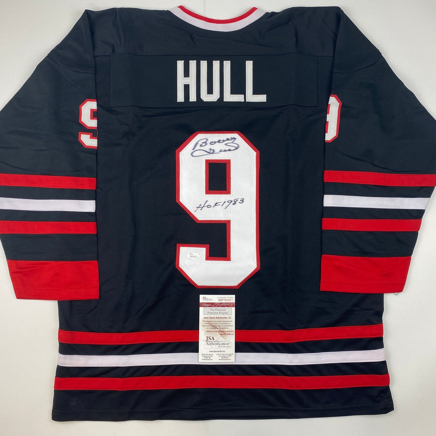 Back view of black hockey jersey with red and white stripes, number 9, "HULL" nameplate, and signed inscription with JSA authentication certificate attached.