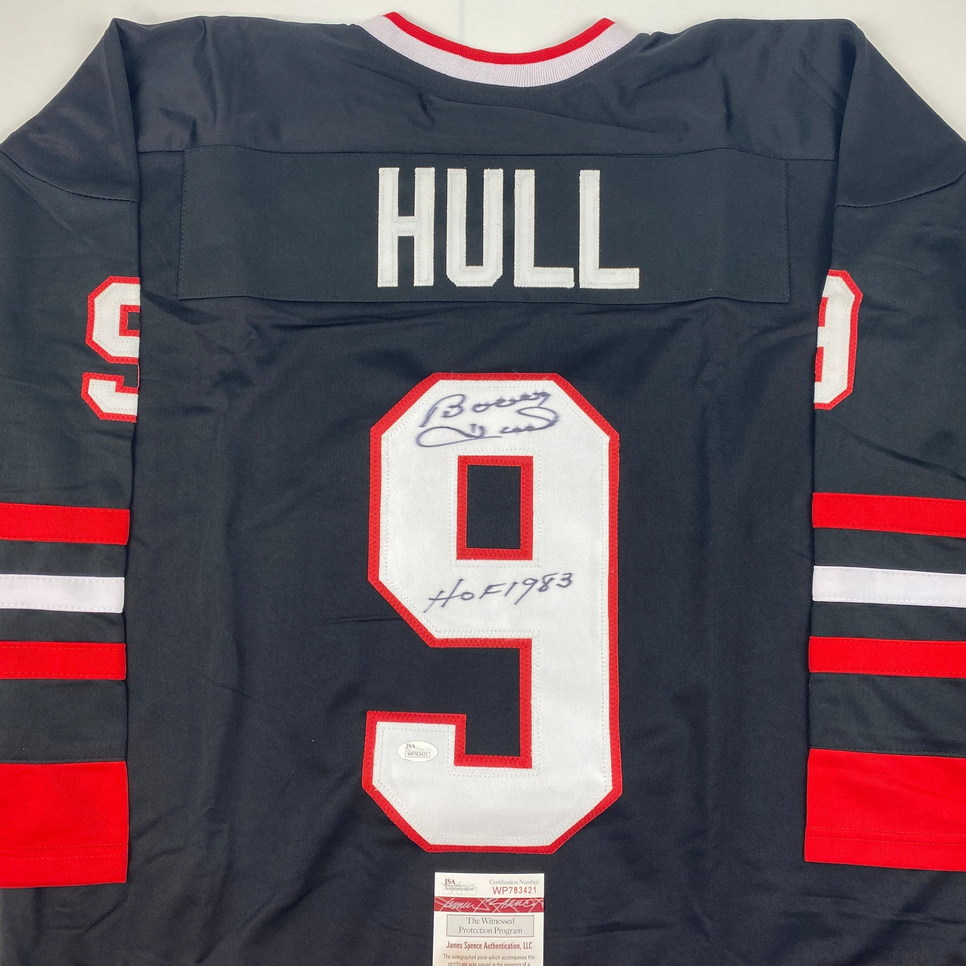 Back view of dark navy hockey jersey with red and white stripes, number 9, "HULL" nameplate, and blue autograph with "HOF 1983" inscription.