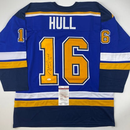 Signed Brett Hull St. Louis Blues jersey displaying number 16 in blue with gold and white trim, flat lay view showing back with JSA authentication certificate.