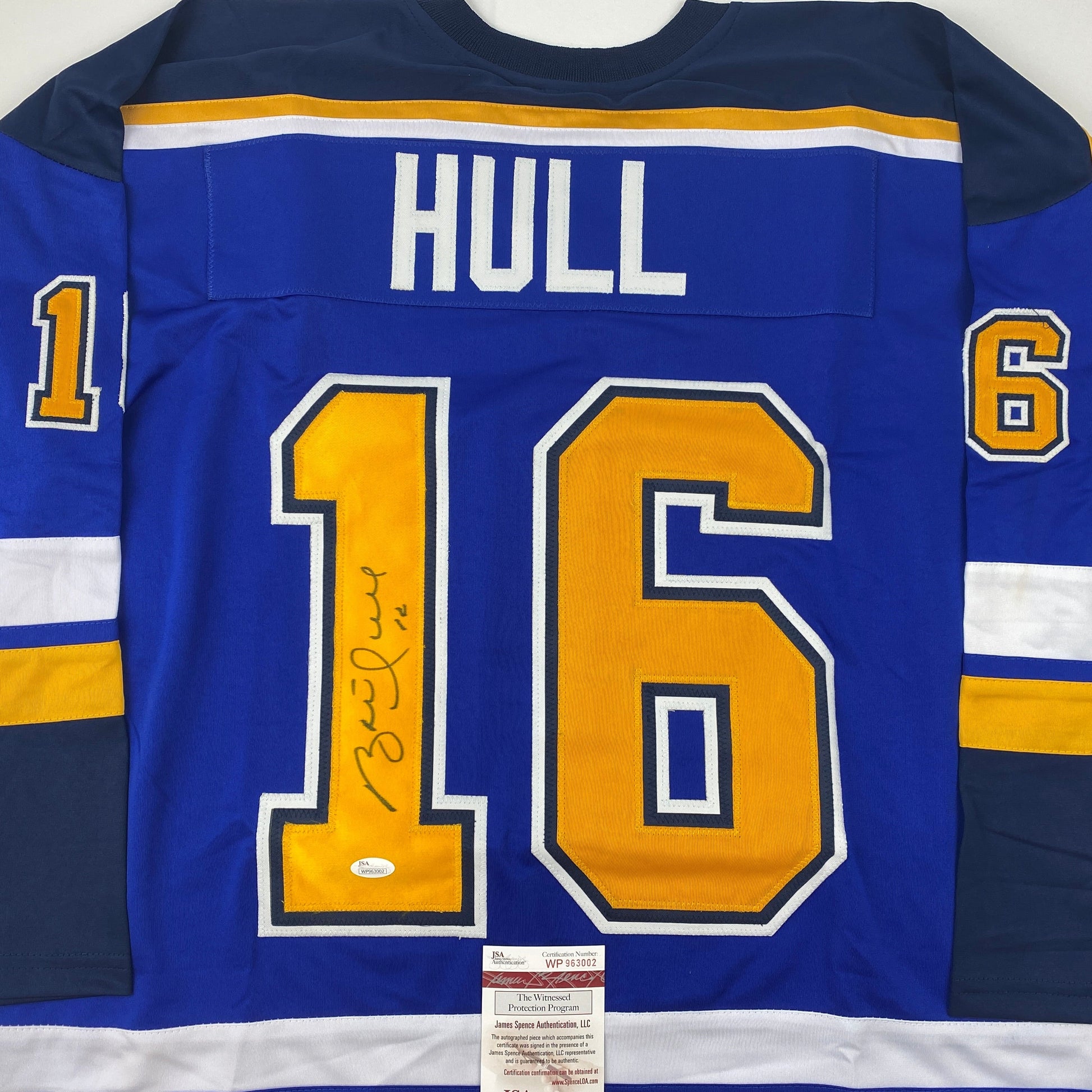 Front view of blue St. Louis Blues hockey jersey number 16 with "HULL" nameplate and autographed signature, displaying yellow and white striping with JSA authentication label.