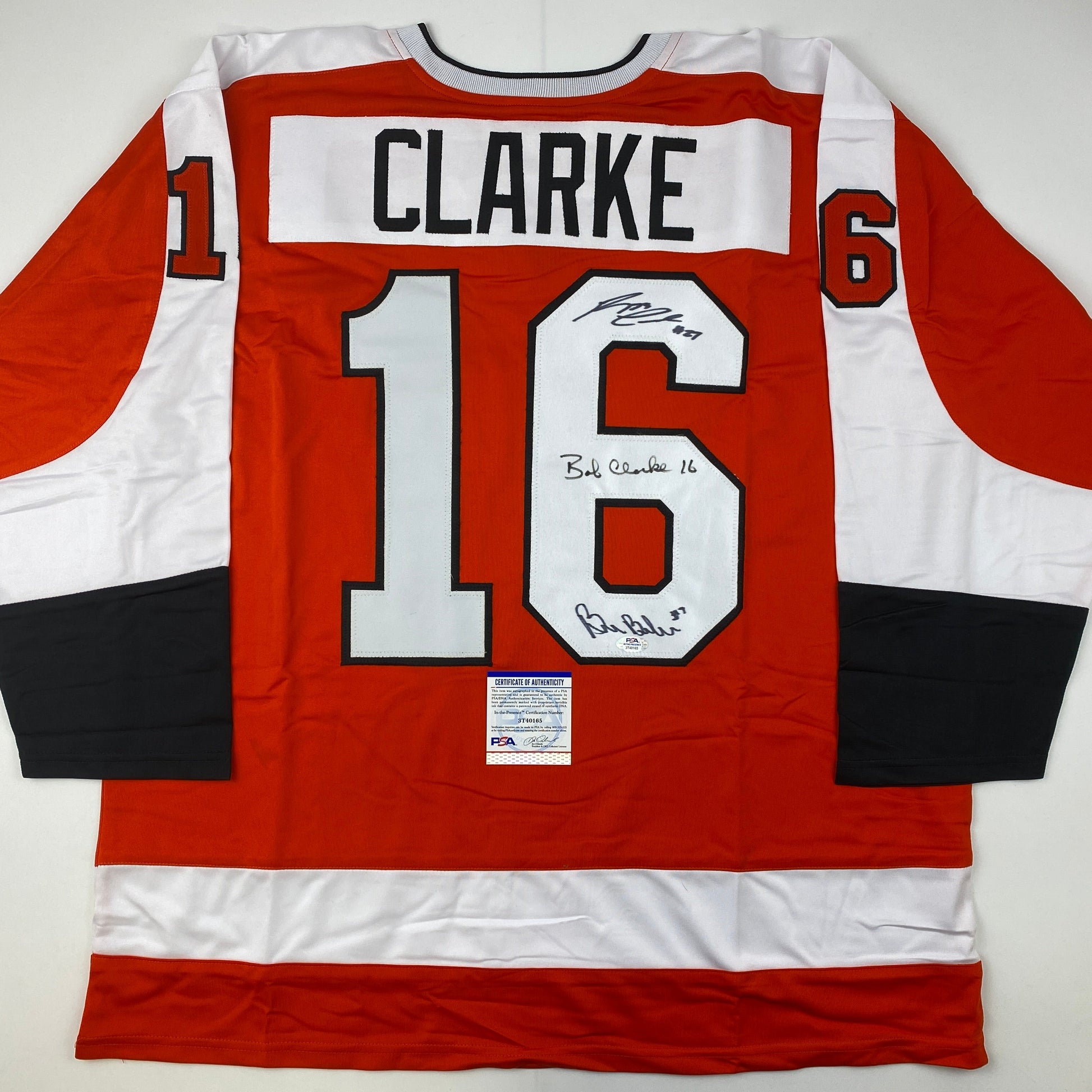 Back view of red and white hockey jersey number 16 with "CLARKE" nameplate and multiple autographs, PSA authentication certificate visible.