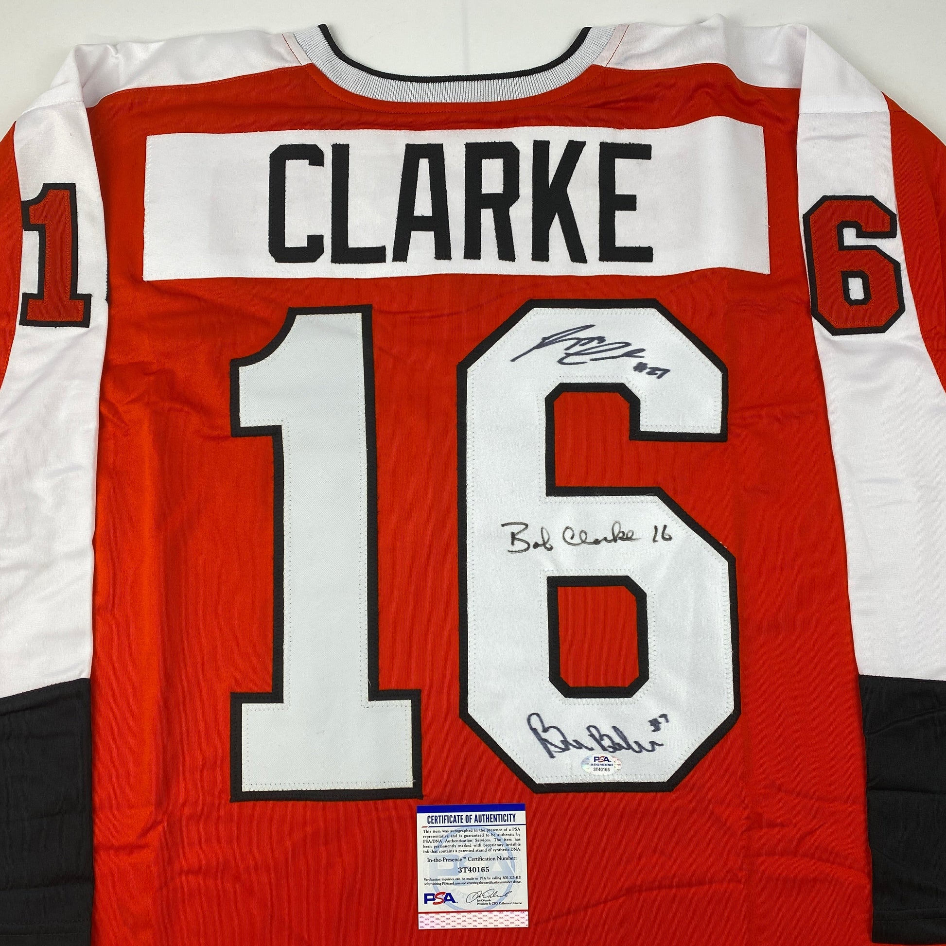 Red and white Philadelphia Flyers hockey jersey number 16 with "CLARKE" nameplate and multiple autographs, displayed flat with PSA authentication certificate visible.