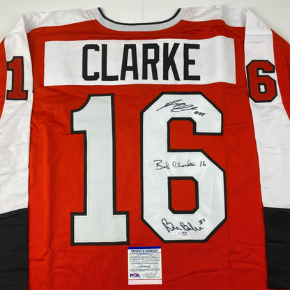 Red and white Philadelphia Flyers hockey jersey number 16 with "CLARKE" nameplate and multiple autographs, displayed flat with PSA authentication certificate visible.