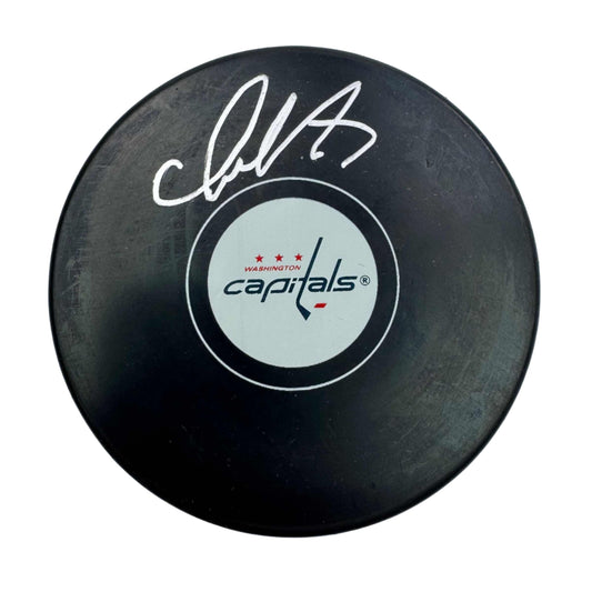 Black hockey puck with white signature and Washington Capitals logo on light circular center label, top-down view.