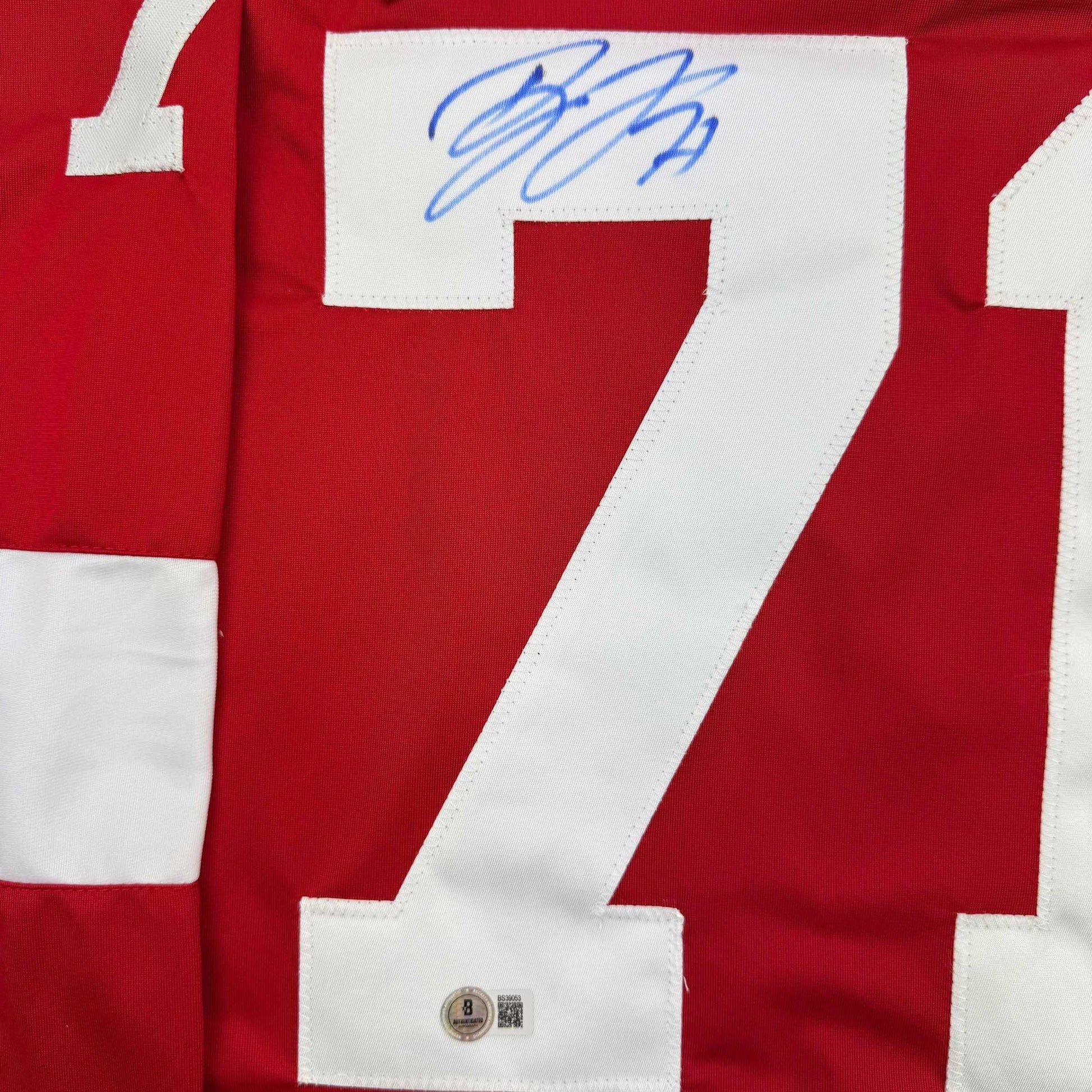 Close-up of red hockey jersey number 7 with blue signature and Beckett authentication hologram visible on white sleeve patch.