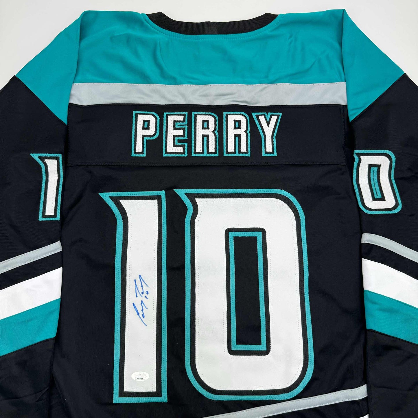 Flat lay of black, turquoise, and white hockey jersey displaying "PERRY" nameplate and number 10 with blue autograph on front.