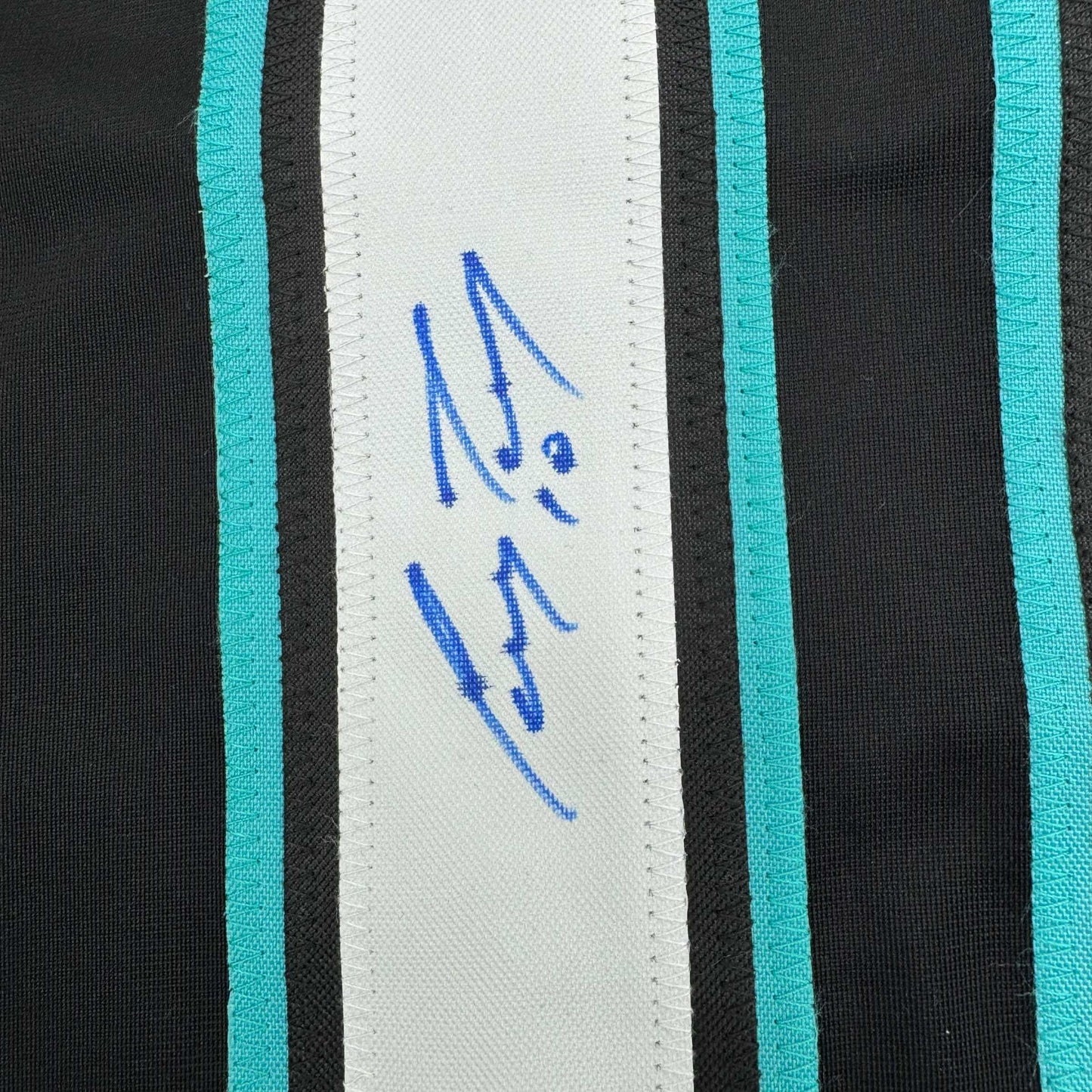 Close-up of blue autograph signature on white stripe with turquoise and black horizontal stripes on hockey jersey.