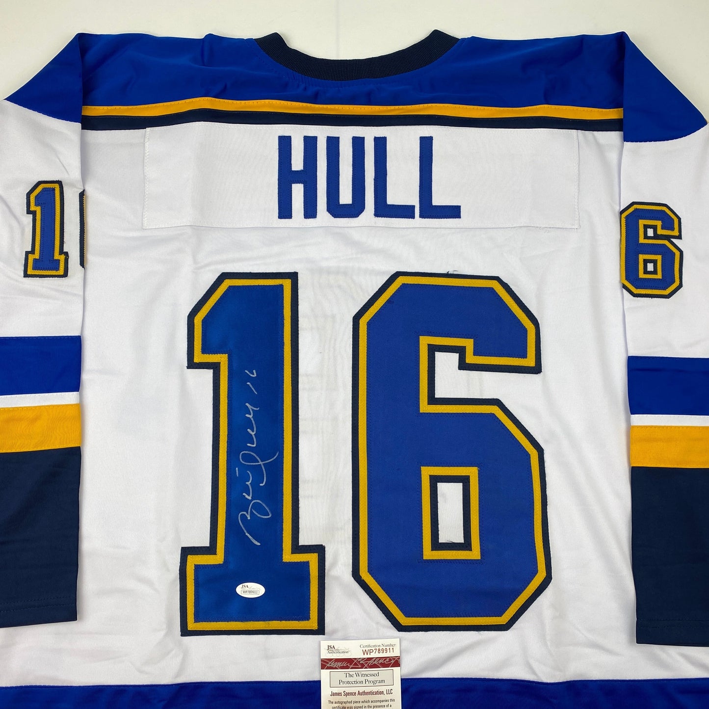 Back view of white hockey jersey with blue and gold stripes, HULL printed across shoulders, large signed number 16, JSA authentication certificate visible at bottom.