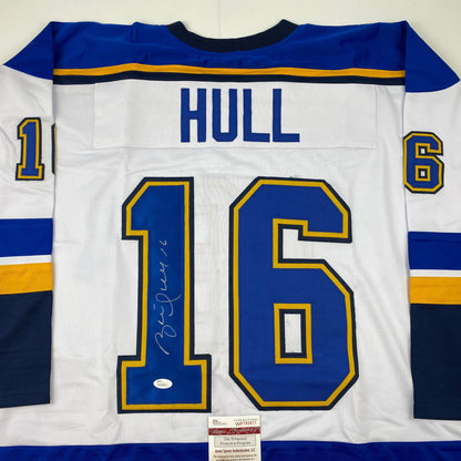 Back view of white hockey jersey with blue and gold stripes, HULL printed across shoulders, large signed number 16, JSA authentication certificate visible at bottom.