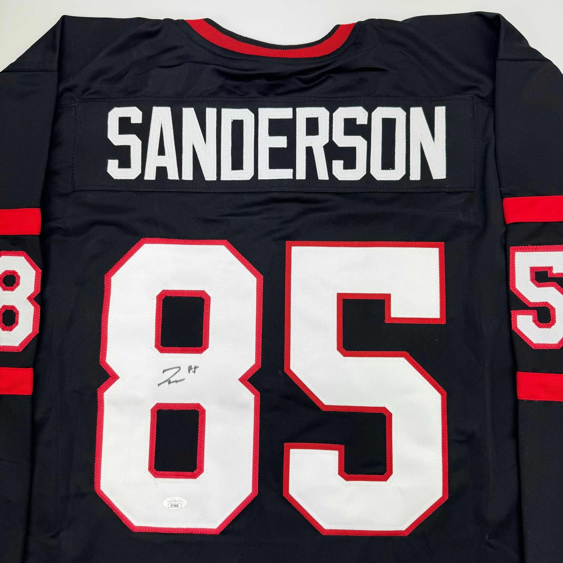 Back view of black hockey jersey with red and white number 85, "SANDERSON" nameplate, and red neckline trim.