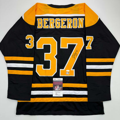 Black and gold Boston Bruins hockey jersey number 37 with "BERGERON" name and autograph, displayed flat with JSA authentication certificate visible.