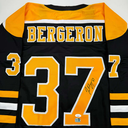 Flat lay of black Boston Bruins jersey with gold and white number 37, "BERGERON" nameplate, and handwritten signature on the orange number.