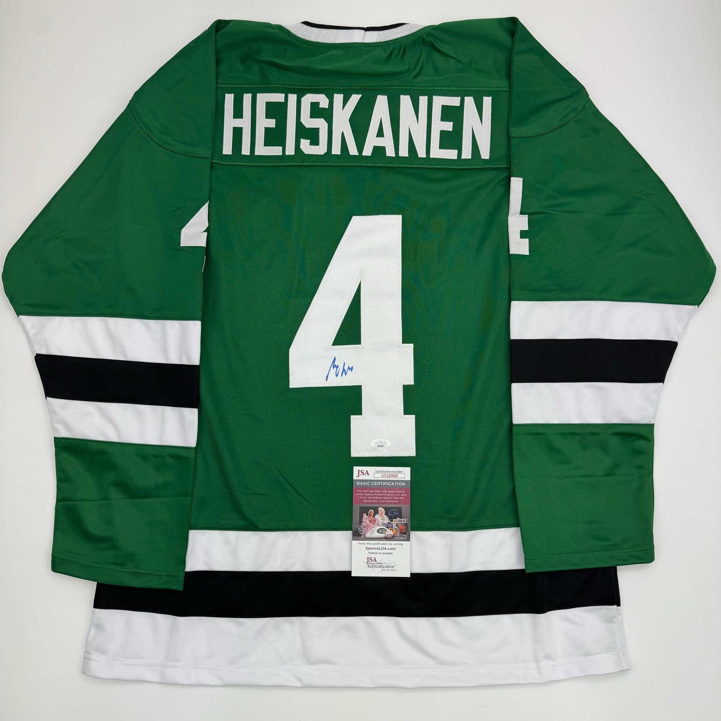 Back view of green hockey jersey with white number 4, "HEISKANEN" nameplate, white and black stripes on sleeves, and JSA authentication certificate attached.