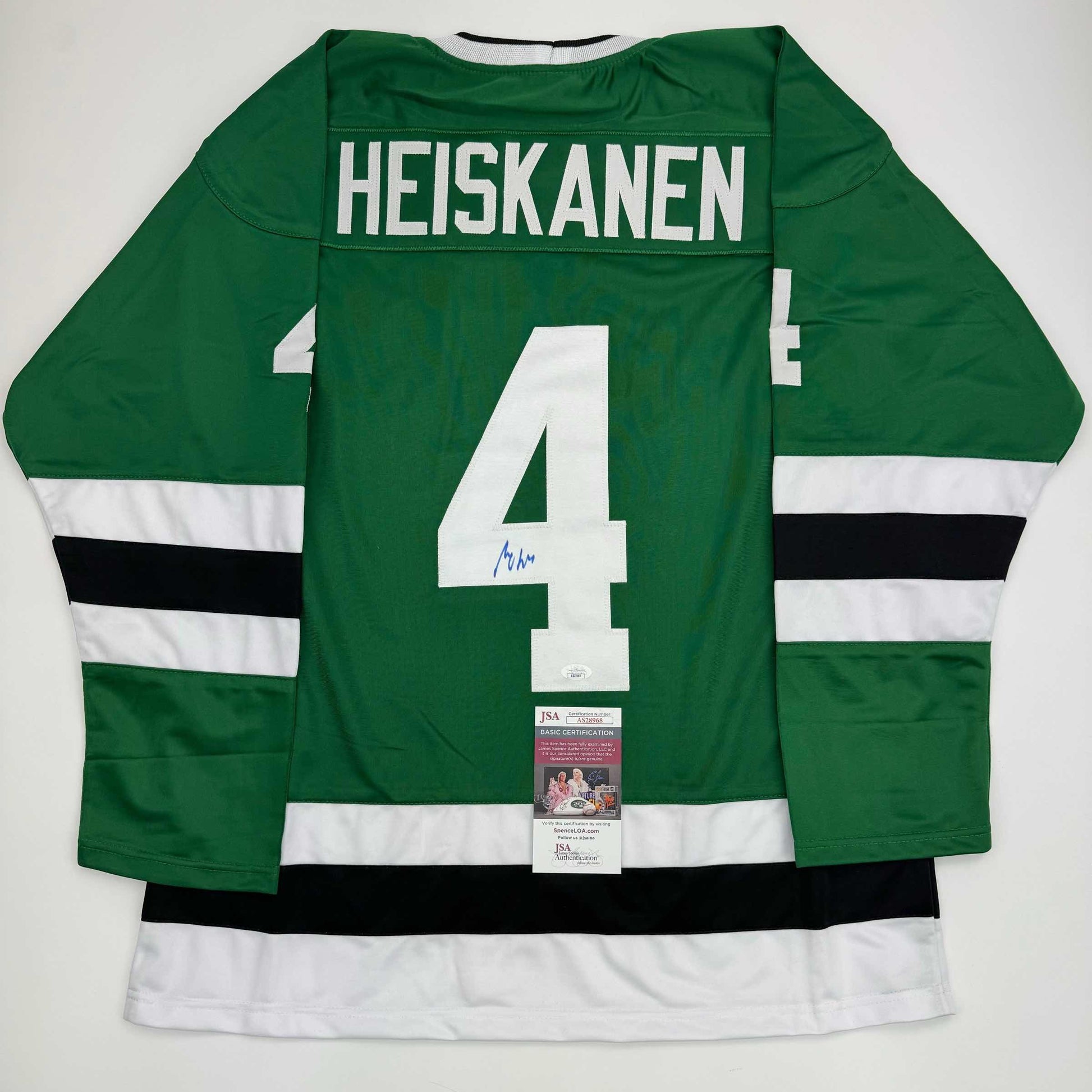 Back view of green hockey jersey with white number 4, "HEISKANEN" nameplate, white and black stripes on sleeves, and JSA authentication certificate attached.