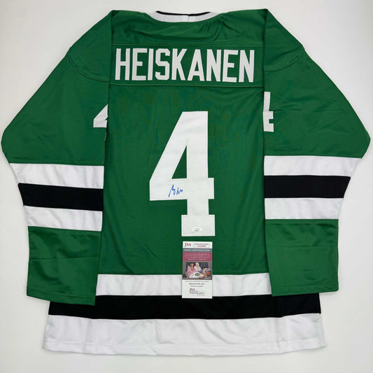 Back view of green hockey jersey with white number 4, "HEISKANEN" nameplate, white and black stripes on sleeves, and JSA authentication certificate attached.