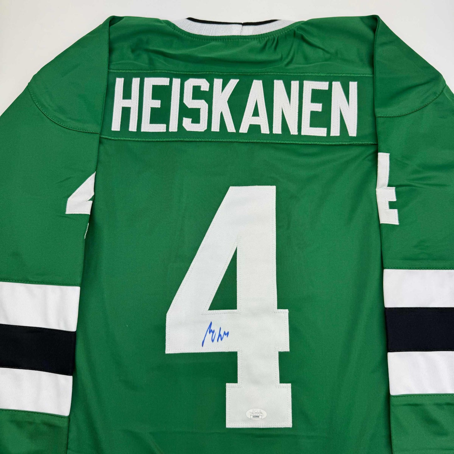 Back view of green hockey jersey with white number 4, "HEISKANEN" nameplate, white shoulder stripes, and blue autograph on the number.