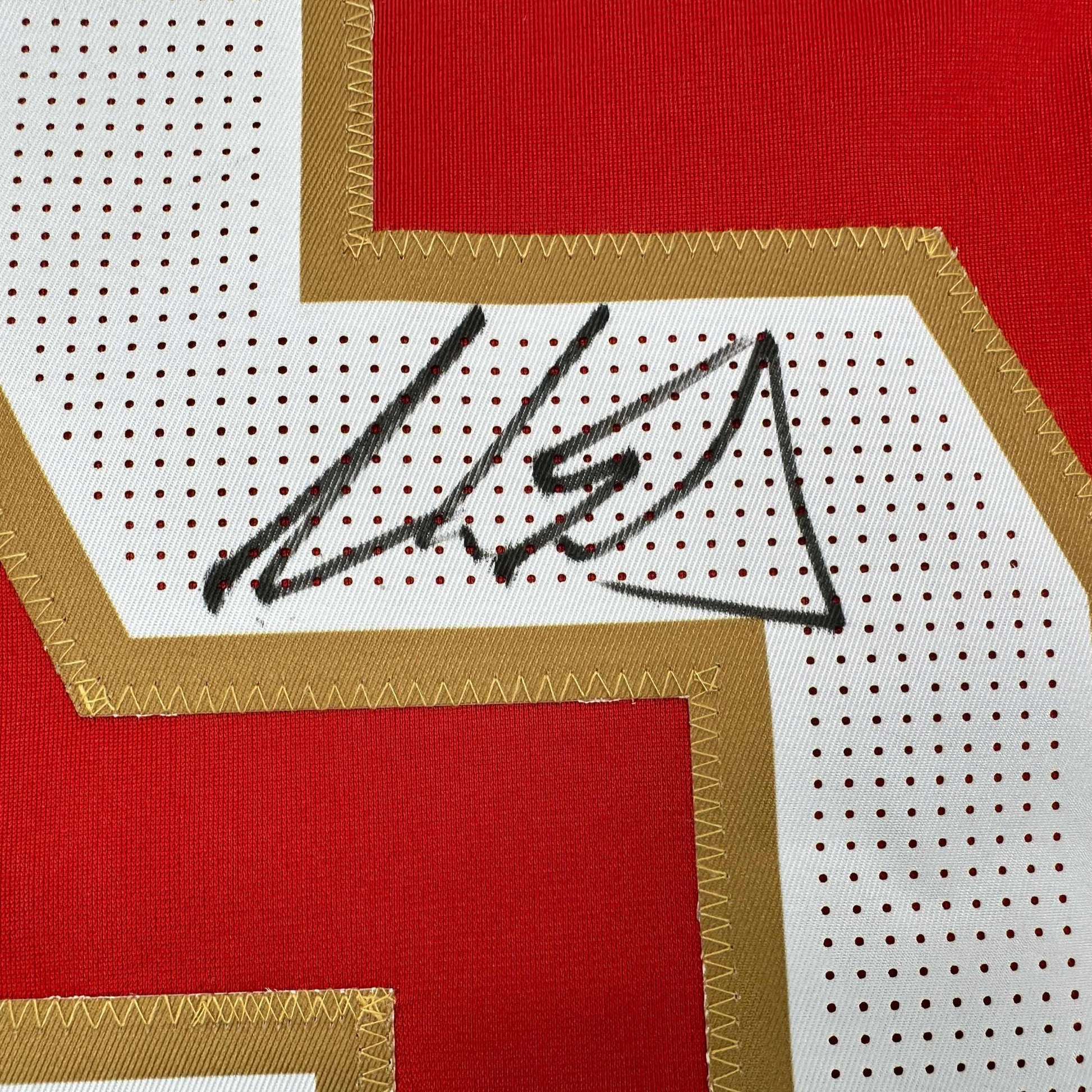 Close-up of black handwritten signature on white mesh panel with red and tan fabric border of hockey jersey.