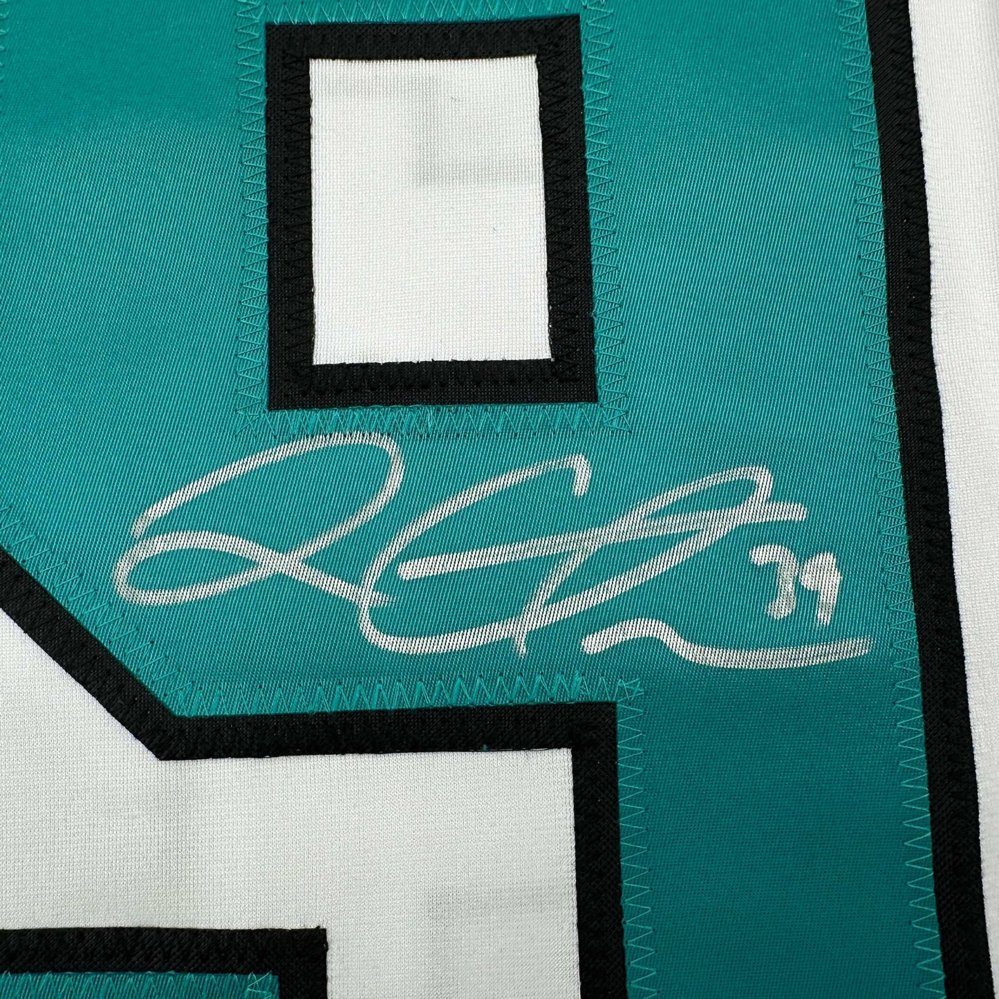 Close-up of teal and white hockey jersey showing cursive white signature on teal fabric with black trim.