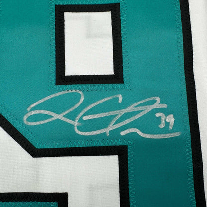 Close-up of teal and white hockey jersey showing cursive white signature on teal fabric with black trim.