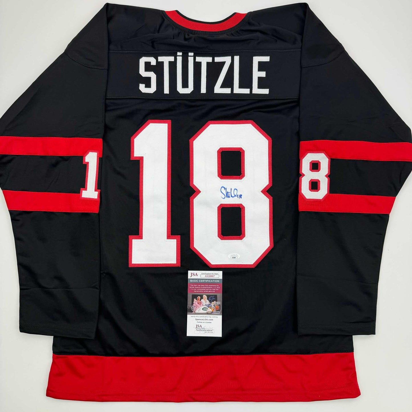 Back view of black hockey jersey with red horizontal stripes, white number 18, "STUTZLE" nameplate, and JSA authentication certificate attached.