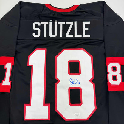 Back view of black hockey jersey with red and white number 18, "STUTZLE" nameplate, red neckline trim, and blue autograph on chest.