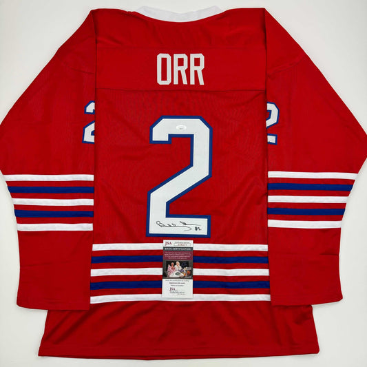 Red hockey jersey with "ORR" and number 2 displayed, showing back view with JSA authentication certificate attached.