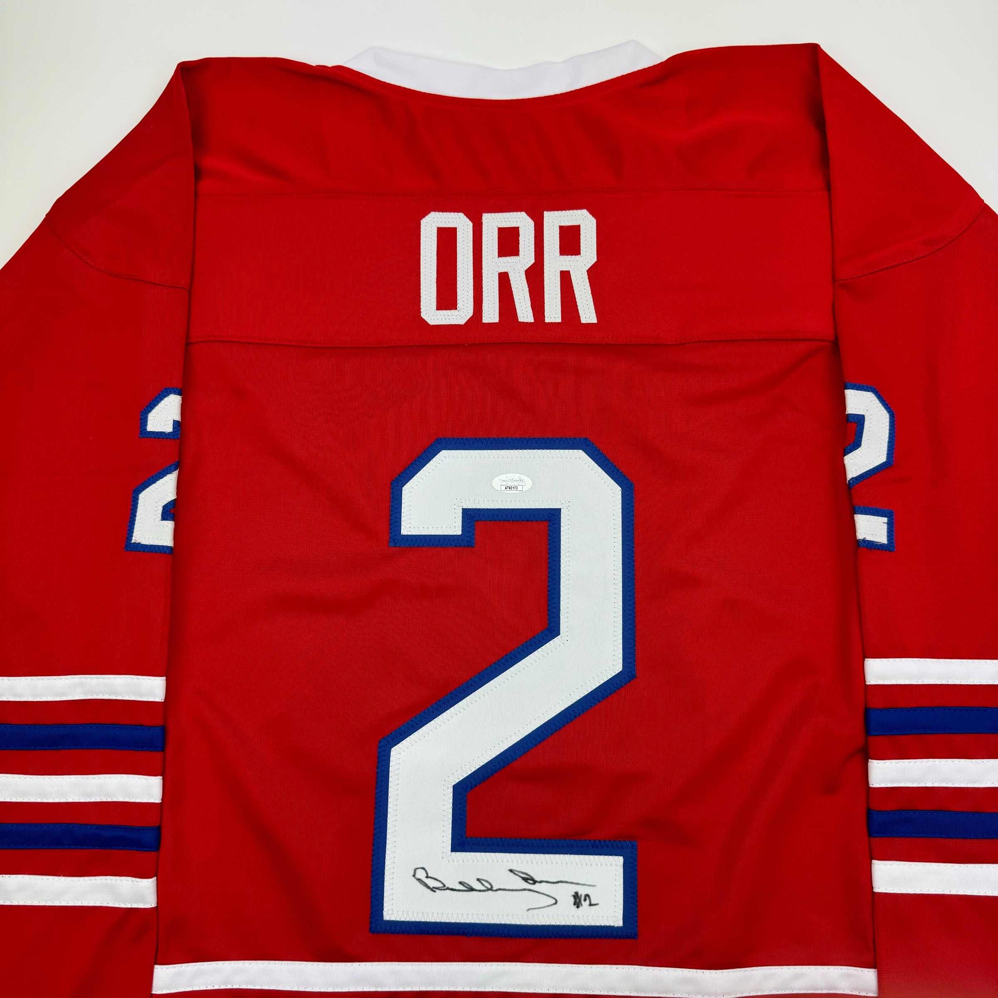Back view of red hockey jersey with "ORR" and number "2" in white and blue, featuring horizontal striped sleeves and handwritten signature on the number.