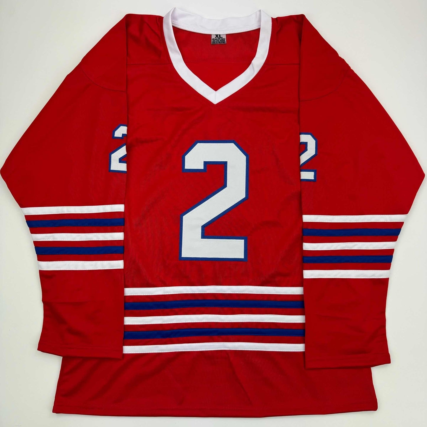 Red hockey jersey with white v-neck collar, large white number 2 with blue outline, red-white-blue striped sleeves and waistband, displayed flat on white background.