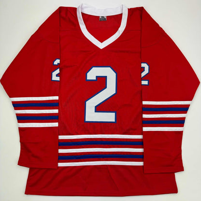 Red hockey jersey with white v-neck collar, large white number 2 with blue outline, red-white-blue striped sleeves and waistband, displayed flat on white background.