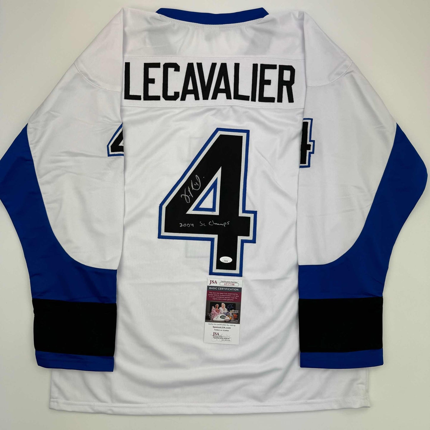 Back view of white hockey jersey with blue and black sleeves, number 4, "LECAVALIER" nameplate, autographed with "2004 SC Champs" inscription, and JSA certification label attached.