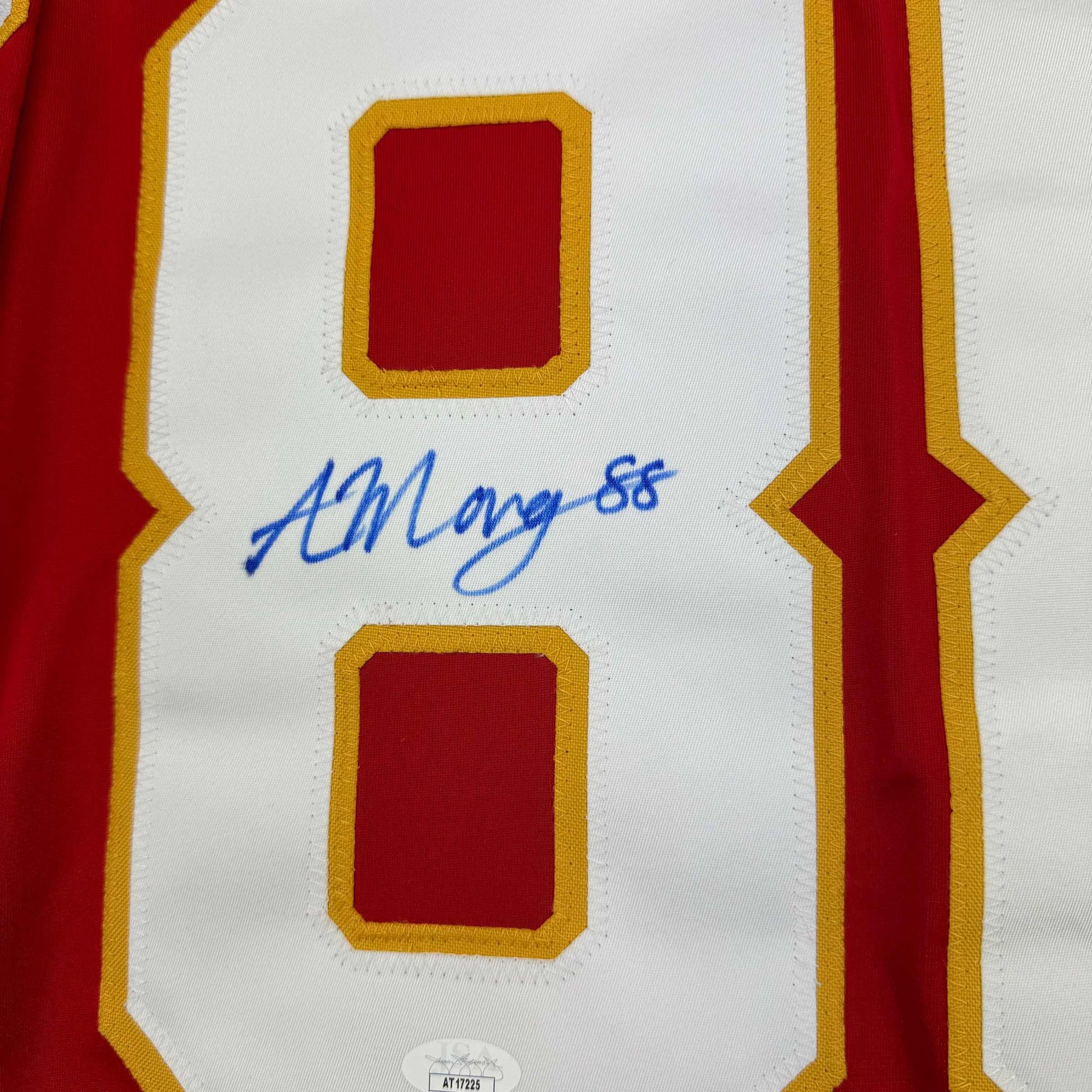 Close-up of white hockey jersey front panel with red and gold trim showing blue autograph "ALMangiapane 88" and JSA authentication label.