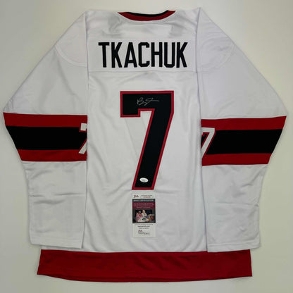 White hockey jersey with "TKACHUK" and number 7 displayed flat, showing red and black striped sleeves and JSA certification label.