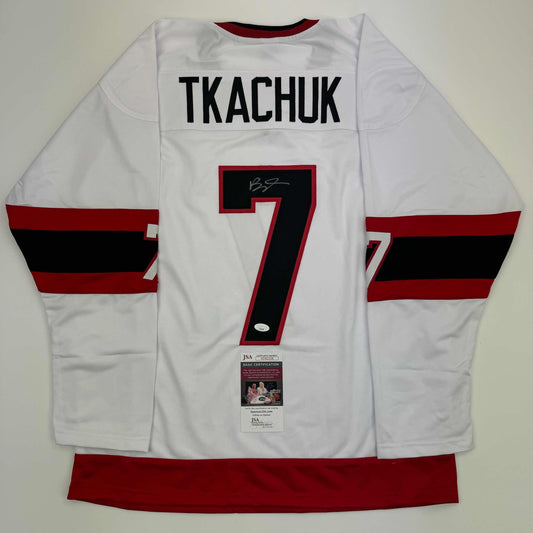 White hockey jersey with "TKACHUK" and number 7 displayed flat, showing red and black striped sleeves and JSA certification label.