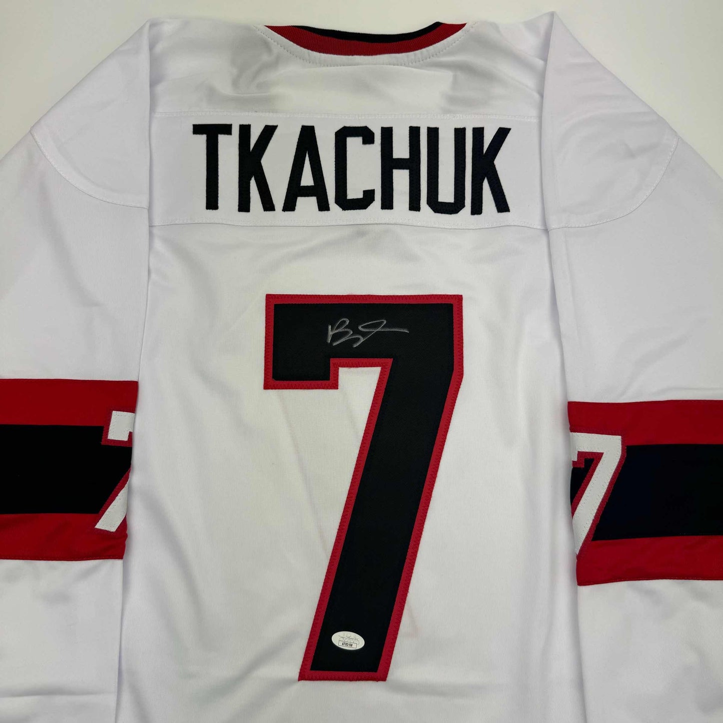 Front view of white hockey jersey with "TKACHUK" nameplate and number 7 in red and black, featuring signature and JSA authentication sticker.