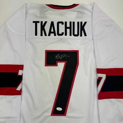 Front view of white hockey jersey with "TKACHUK" nameplate and number 7 in red and black, featuring signature and JSA authentication sticker.