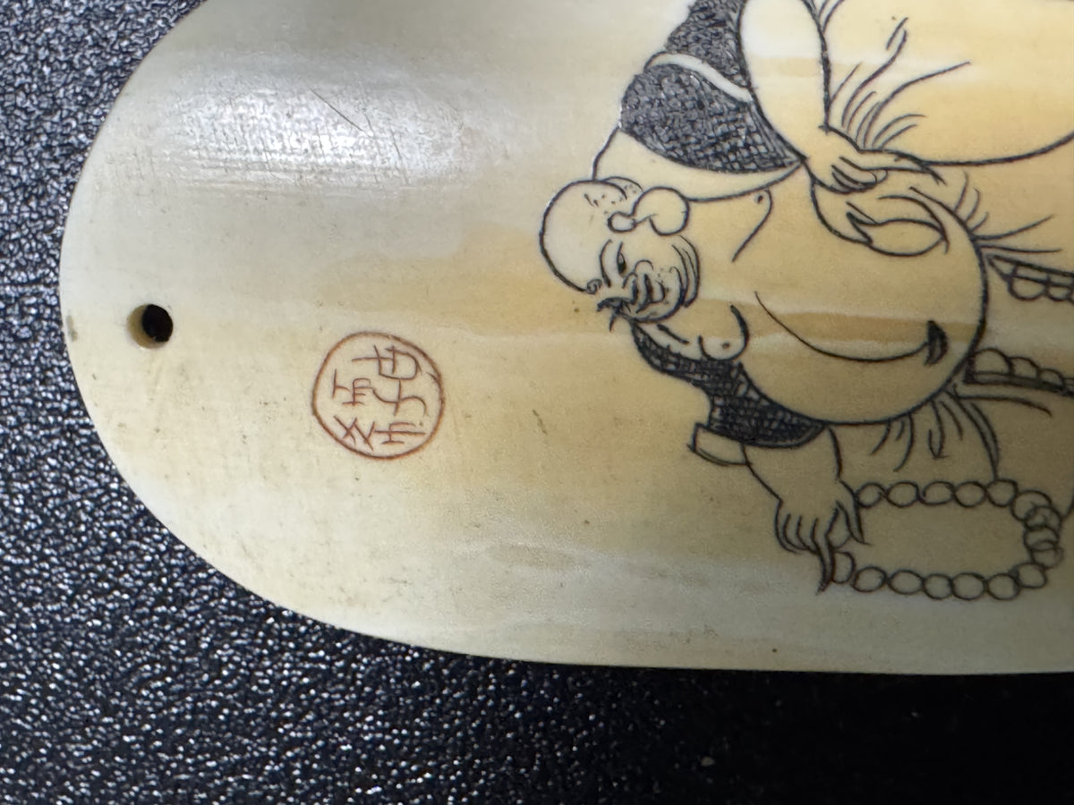 Macro of the front of the ox horn pendant against a dark ground: the round red seal beside the laughing figure's head, its seal-script strokes worn but visible.