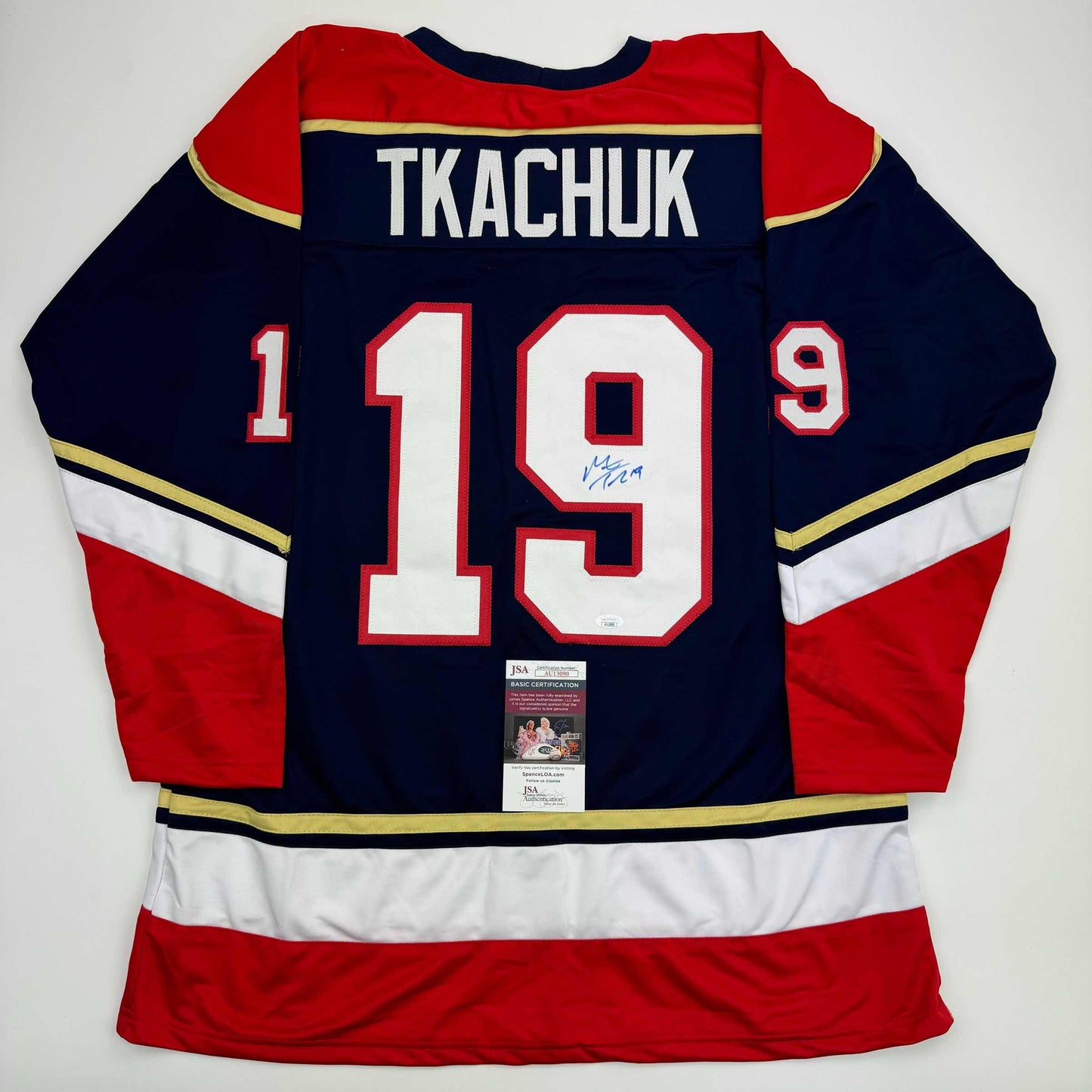 Back view of navy blue hockey jersey with red and white stripes, displaying "TKACHUK" and number "19" with signature and JSA certification label.