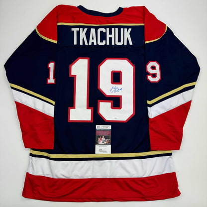 Back view of navy blue hockey jersey with red and white stripes, displaying "TKACHUK" and number "19" with signature and JSA certification label.