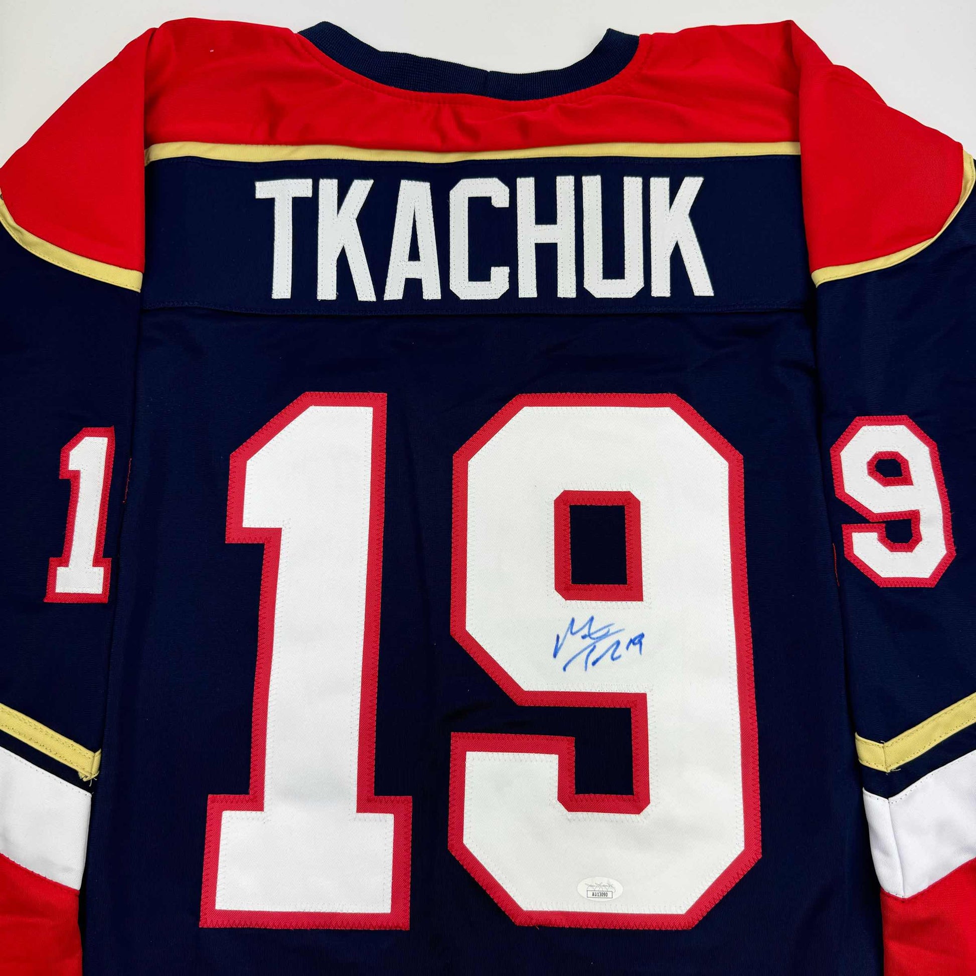 Back view of navy blue hockey jersey with red and gold trim displaying "TKACHUK" and number 19 with blue autograph on white numbering.