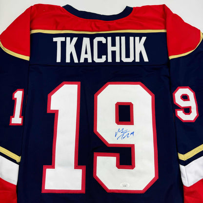 Back view of navy blue hockey jersey with red and gold trim displaying "TKACHUK" and number 19 with blue autograph on white numbering.