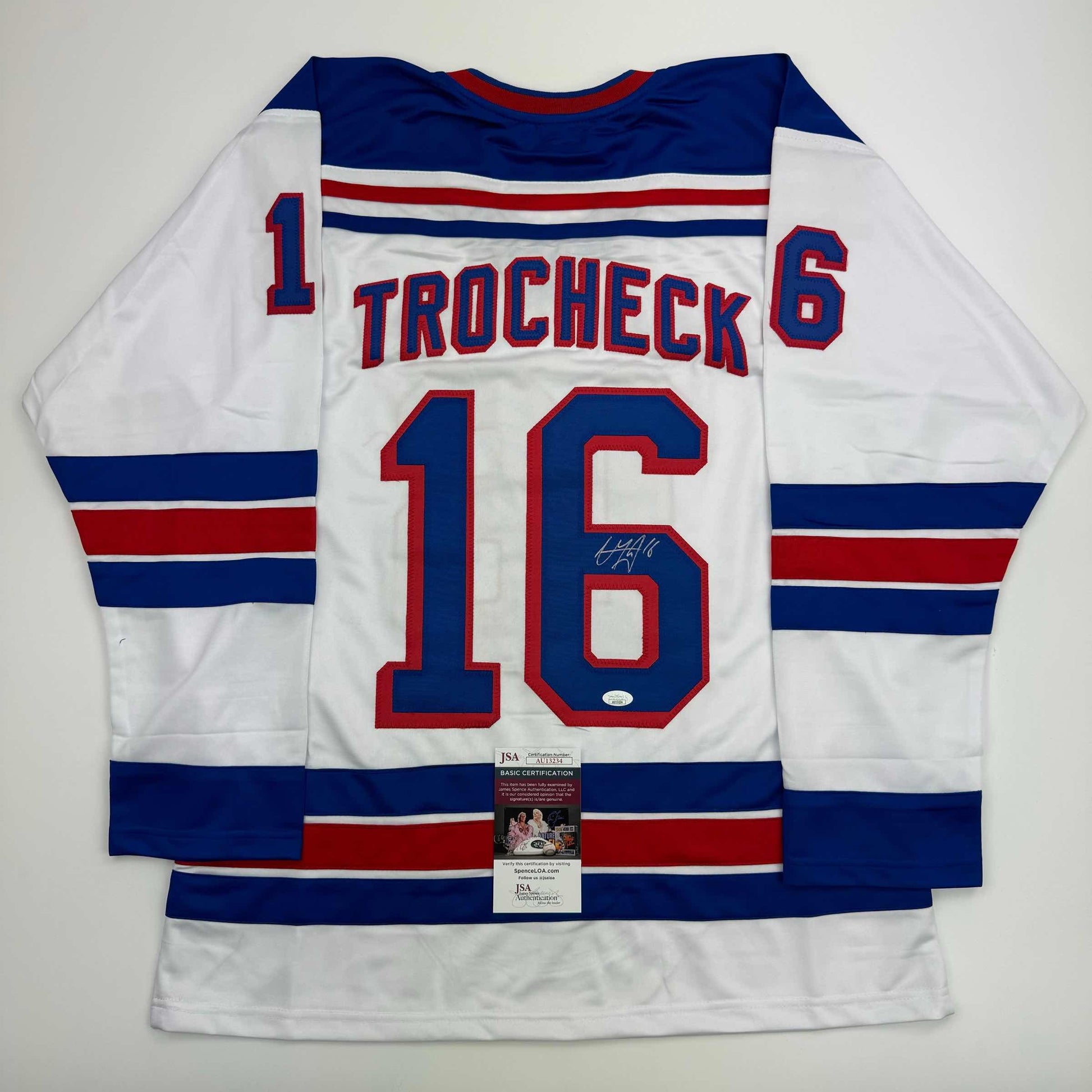 White hockey jersey with navy and red stripes, displaying "TROCHECK 16" and autograph, shown flat with JSA authentication certificate visible.